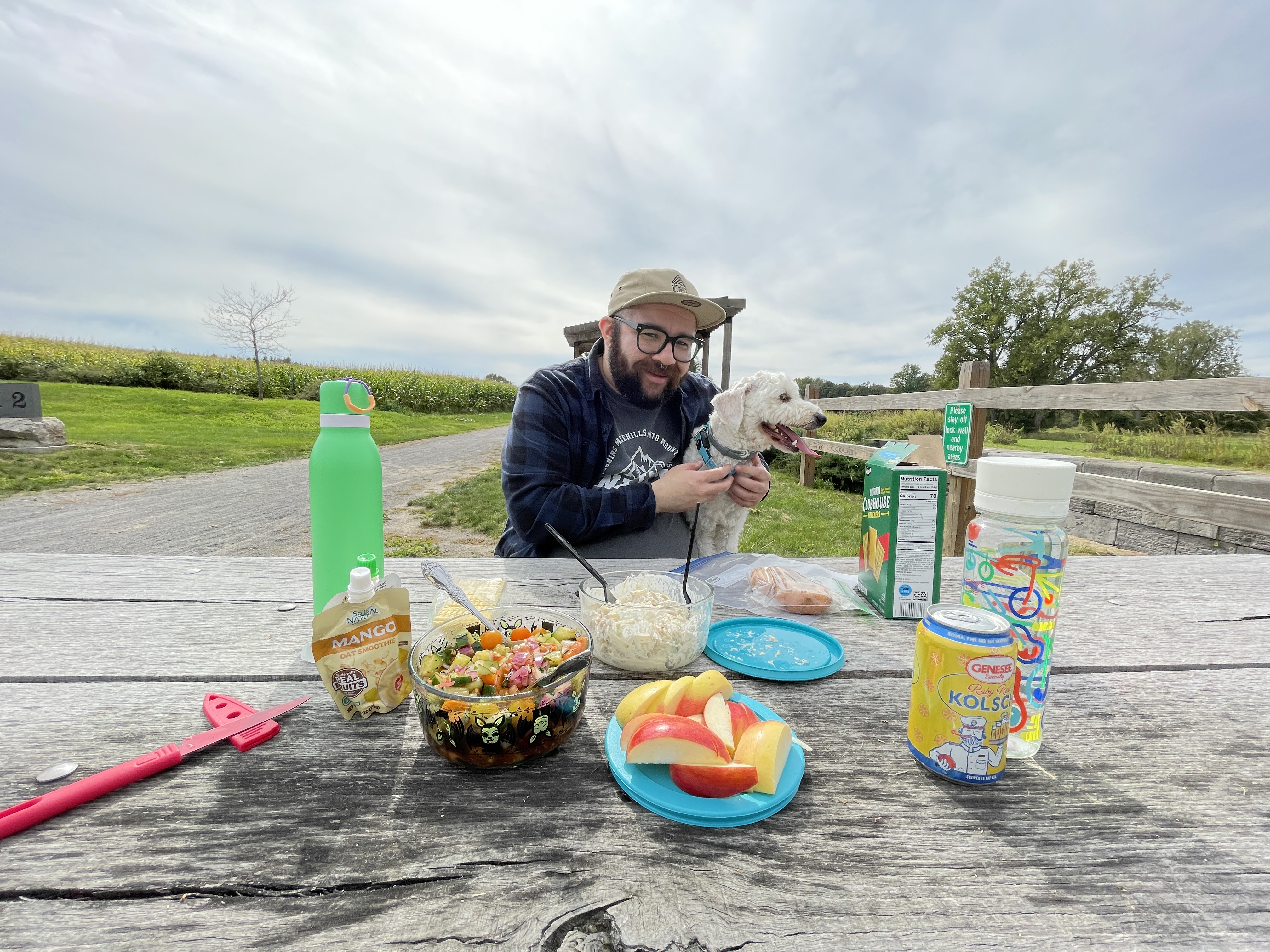 Various foods spread out on a picnic table. A man with a flannel and a hat on smiling at the camera with his arm around a small white poodle