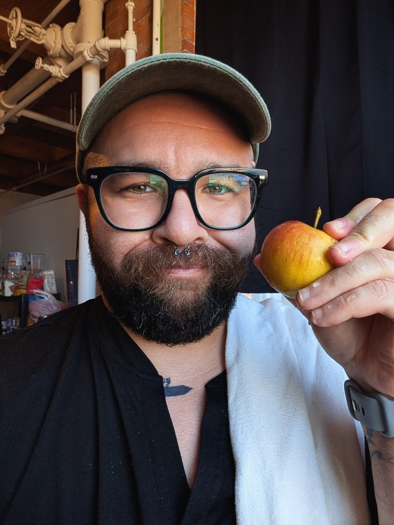 a man with glasses close up wearing a hat. he has a beard. he is holding a tiny red and yellow apple and making a funny face